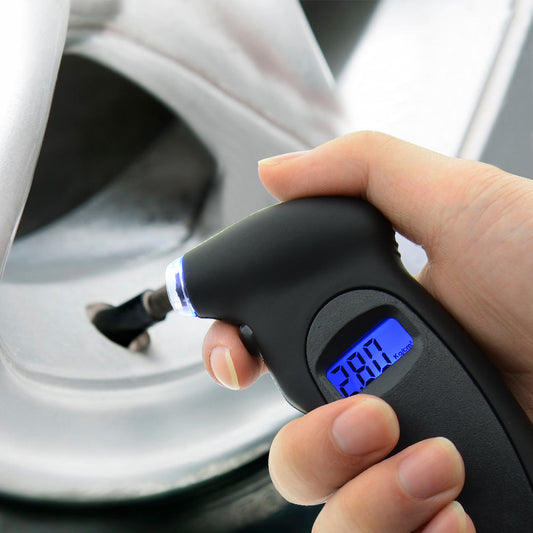 A hand holding a digital tire pressure gauge with a blue LCD screen, against the backdrop of a car tire.